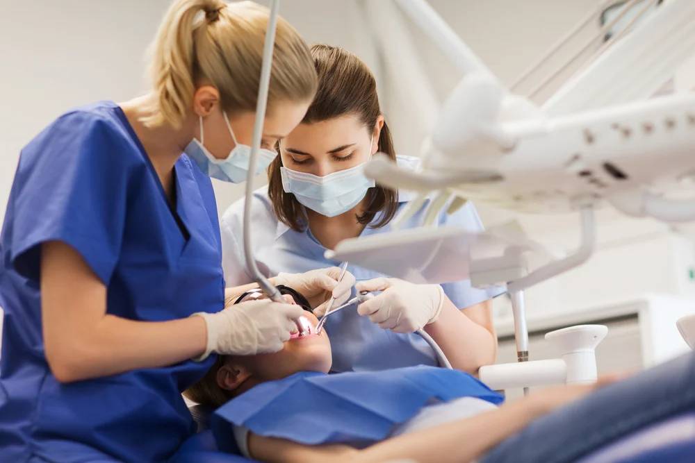 Two dental professionals in blue scrubs and masks work together on a patient in a modern clinical setting